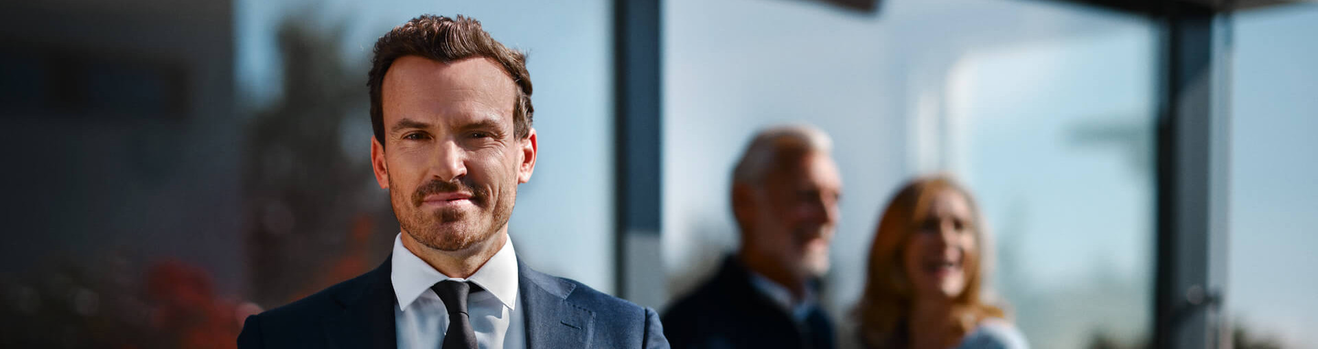 Selbstbewusster Makler sieht in die Kamera mit älterem Ehepaar und Haus im Hintergrund