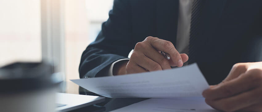 Mann im Anzug sitzt am Tisch mit Stift in der Hand und schaut sich ein Vertrag an