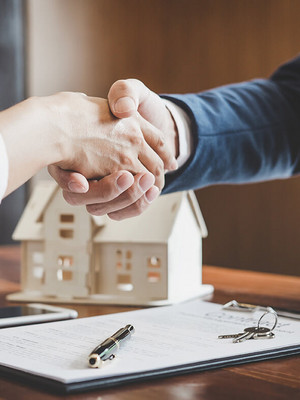 Personen geben sich einen Handschlag mit Modellhaus im Hintergrund