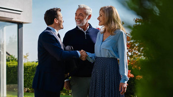 Makler und älteres Paar stehen im Garten und Makler schüttelt die Hand der Frau